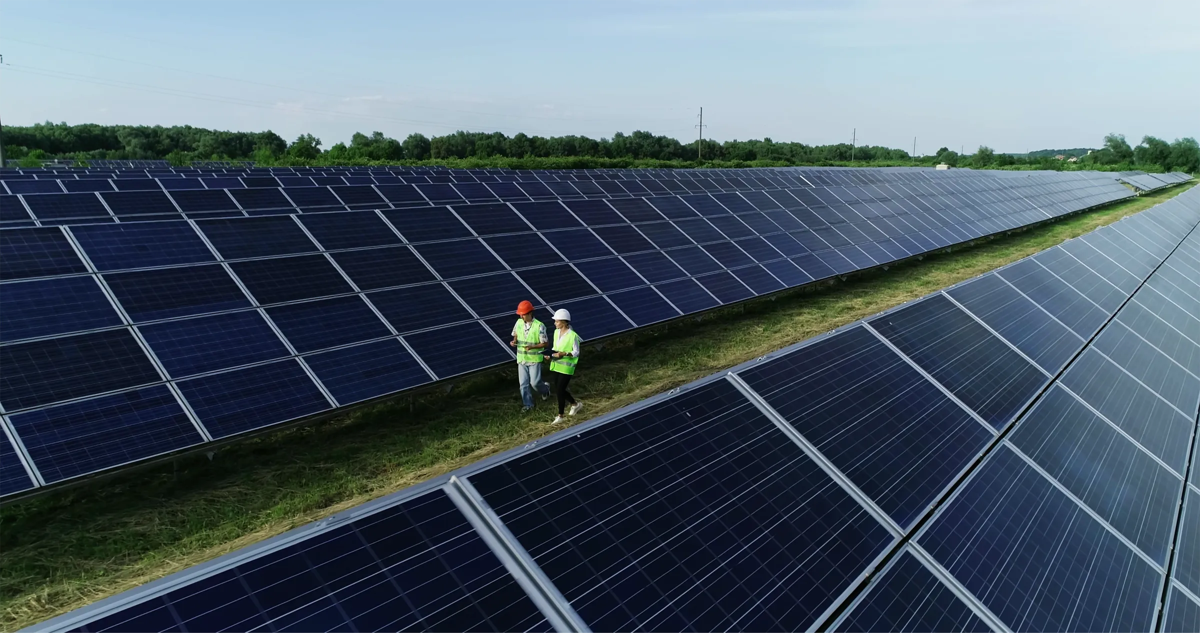 Solar energy technician in a hard hat inspecting a large row of solar panels in a grassy field.
